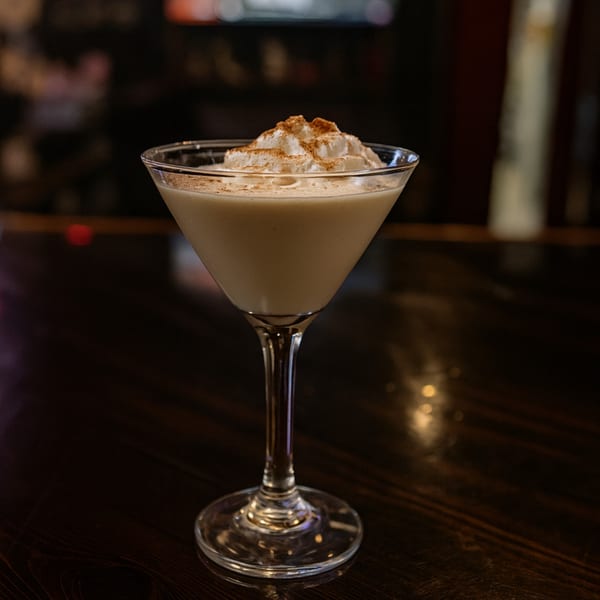 A creamy martini with whipped cream and cinnamon on a dark wooden bar top.