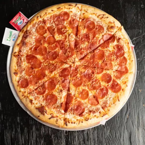 Large, sliced pepperoni pizza on a metal tray with two Lisanti cheese packets on a dark wooden table.