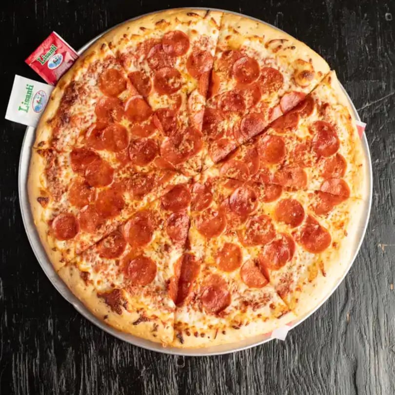 Large, sliced pepperoni pizza on a metal tray with two Lisanti cheese packets on a dark wooden table.