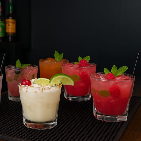 Assortment of five colorful cocktails, garnished with fruit and mint, on a dark bar mat.